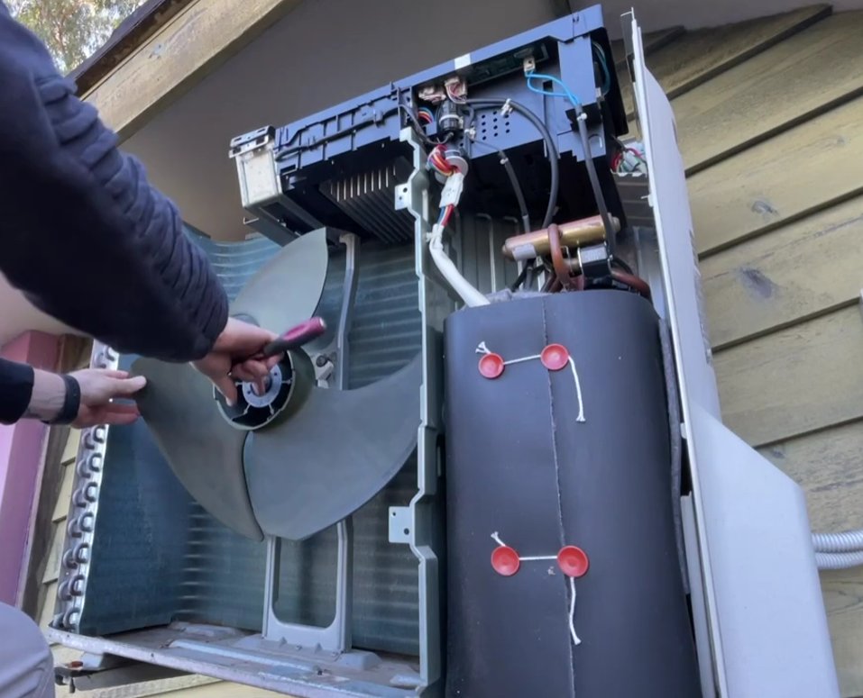 Technician inspecting inside of opened Daikin outdoor unit showing fan blades, motor, wiring and compressor components