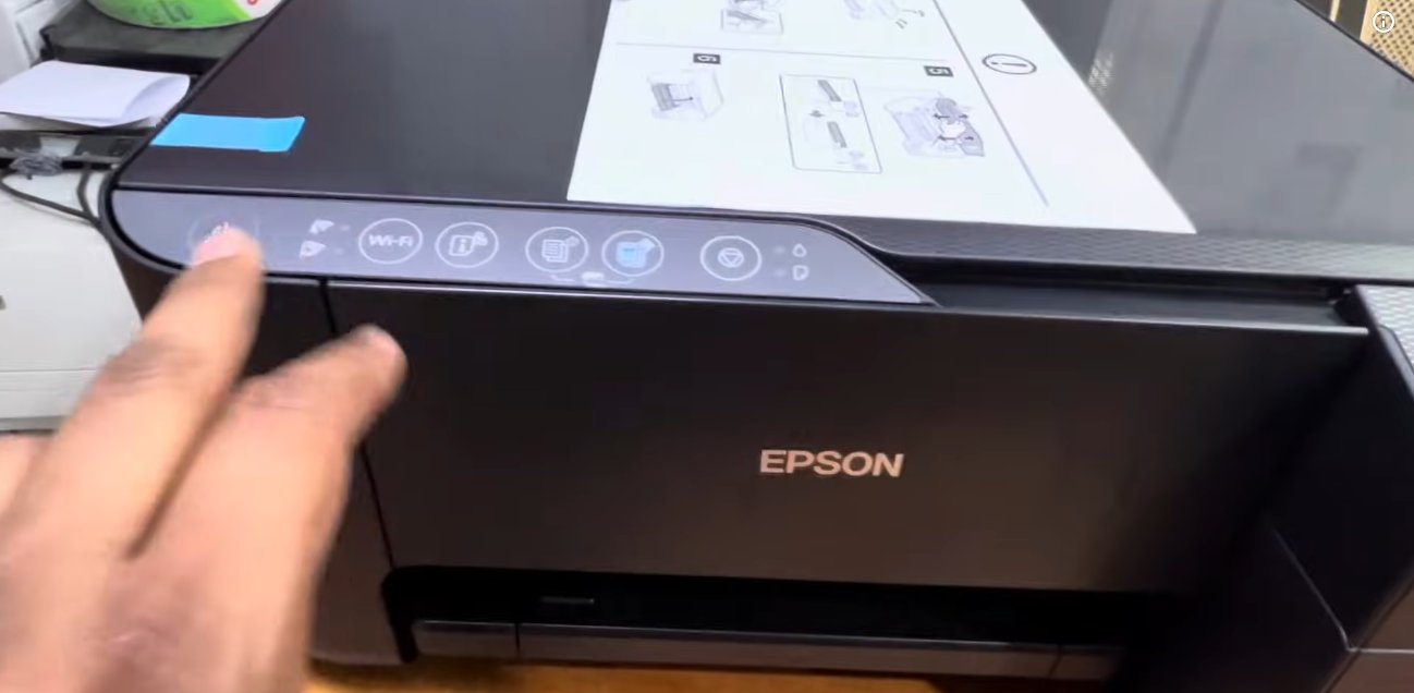 Epson L3250 EcoTank printer viewed from a low angle showing the power button on the far left of the control panel being pressed with the scanner lid open and a printed page visible on the glass