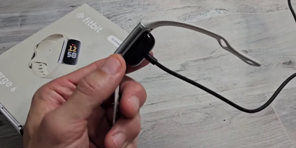 Hand holding a black Fitbit Charge fitness tracker with the magnetic charging cable attached to the back of the device showing the correct cable connection method with the Fitbit retail box visible in the background