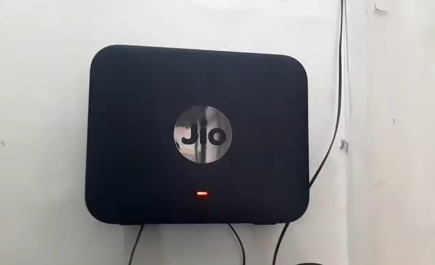 JioFiber ONT device mounted on white wall showing single red blinking LOS indicator light at the bottom of the device with black fibre cables connected