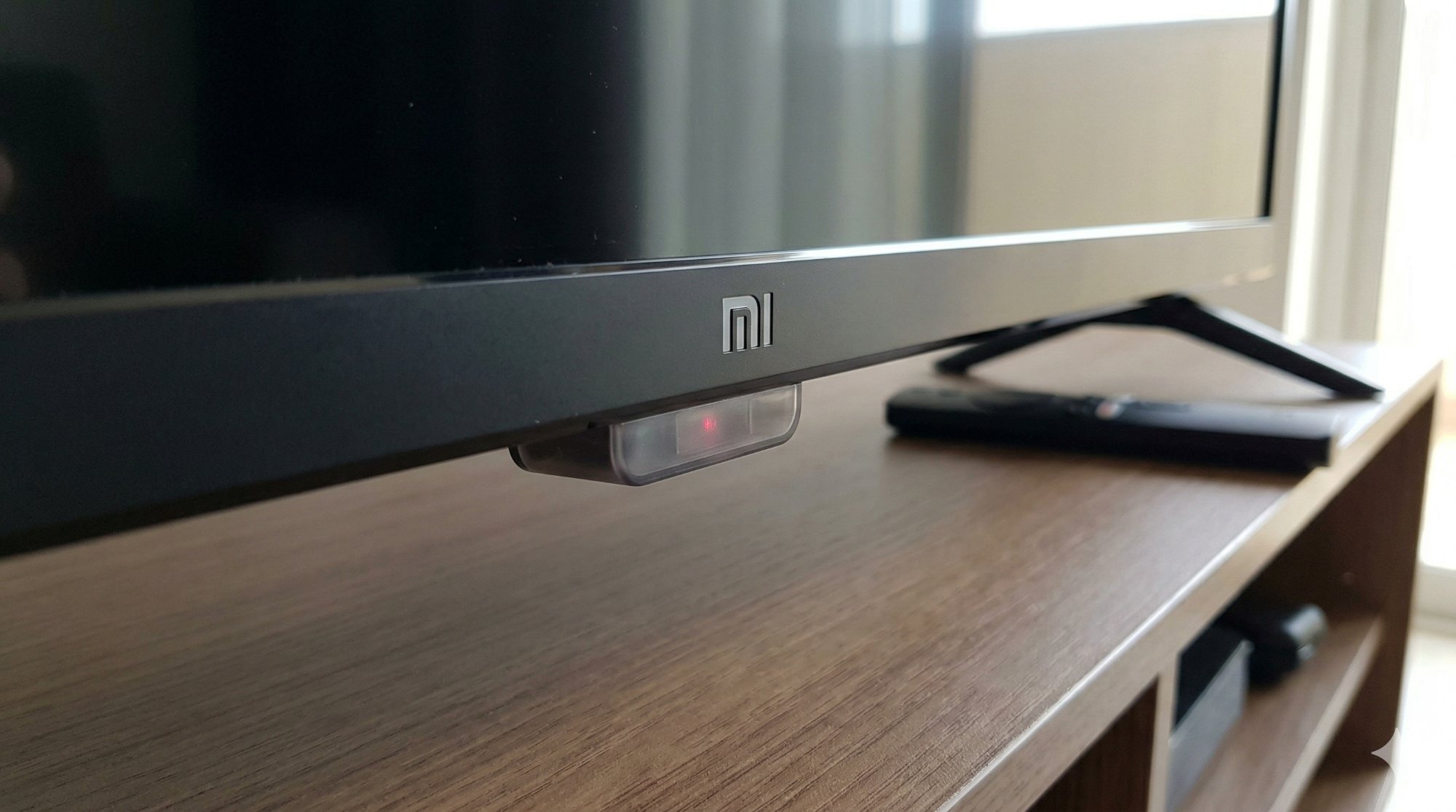 Close-up of Mi TV bottom bezel showing the IR and Bluetooth sensor window with red standby indicator light visible on wooden TV unit