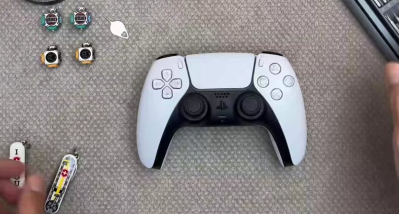 PS5 DualSense controller on grey carpet surface with replacement analogue stick potentiometer modules Hall effect modules and SIM eject tool laid out beside it showing the components needed for Fix 5 potentiometer replacement