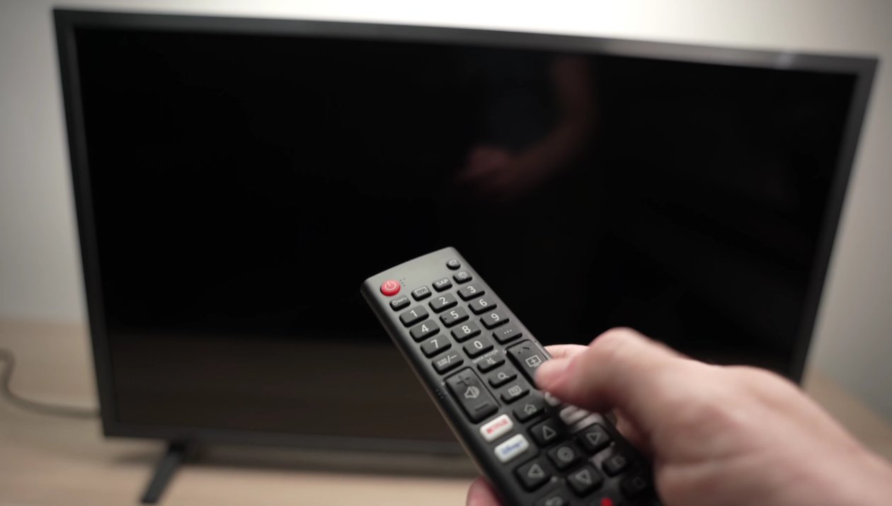 Person holding Samsung TV remote pointing at a black screen Smart TV that shows no image and has the red standby light visible blinking at the bottom edge