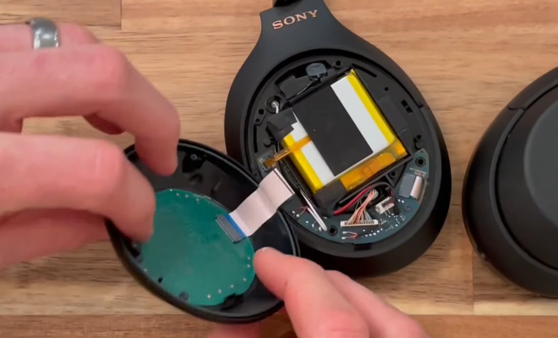 Sony WH-1000XM4 earcup opened showing internal components with the lithium polymer battery pack visible in yellow and white with the green touch sensor board being lifted away from the main PCB showing flex cable connection