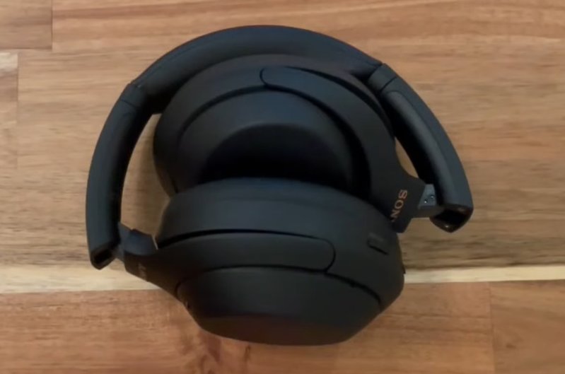 Sony WH-1000XM4 headphones folded flat and viewed from the side on a wooden surface showing the folded compact form and the USB-C charging port location on the left earcup edge