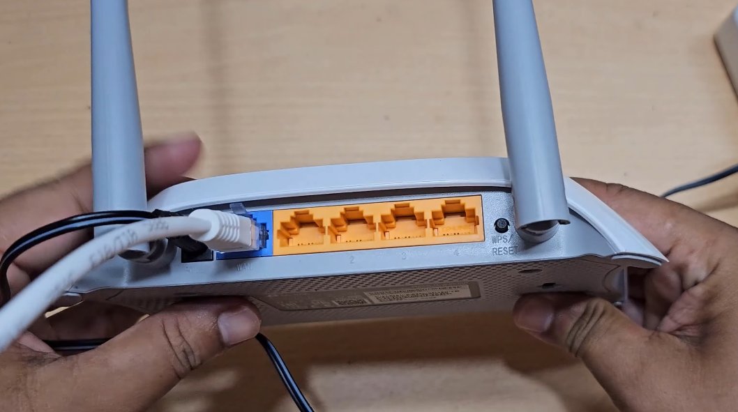 Hands holding the back of a TP-Link router showing the blue WAN port with a white Ethernet cable plugged in and four yellow LAN ports beside it